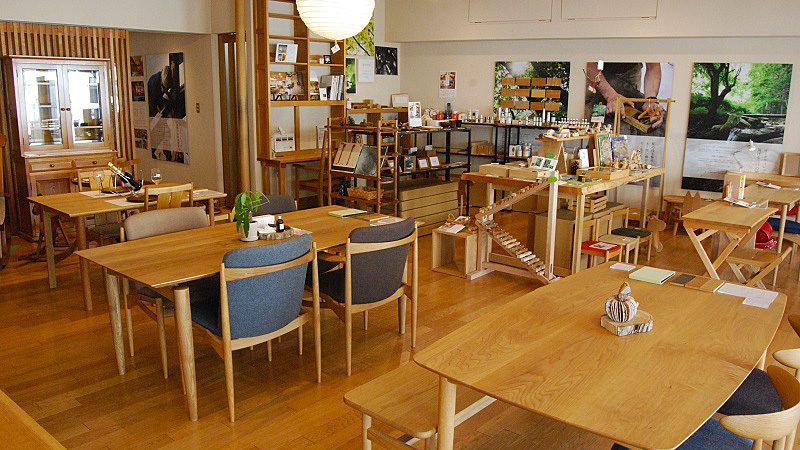 オークヴィレッジ自由が丘の公式店舗ページに掲載された店内写真。木のテーブルと青い座面の椅子が並び、奥の棚に木の小物や文具が陳列され、壁に森の写真が掛かっている
