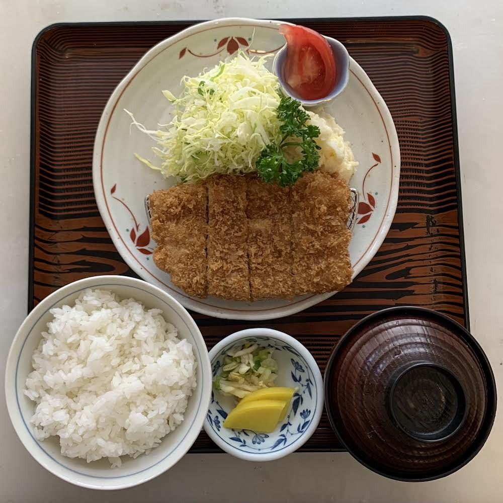 木目の盆に載った定食。絵付けの皿にとんかつと千切りキャベツ、ポテトサラダ、トマト、手前にご飯茶碗、香の物の小鉢、汁椀
