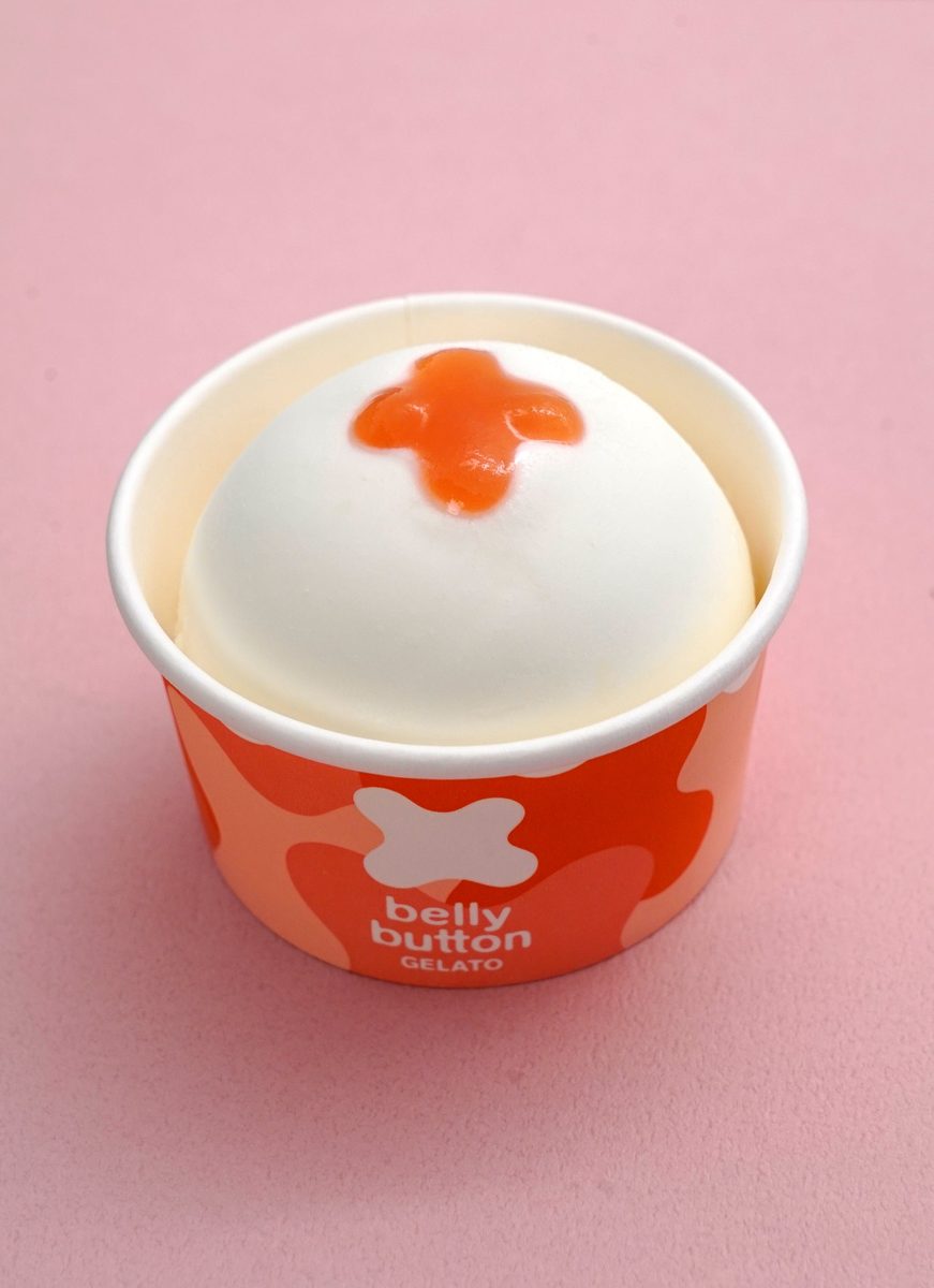 「belly button GELATO」と印刷されたオレンジ色の紙カップに、白いジェラートが丸く盛られ、上にオレンジ色のソースが×印のように落ちている