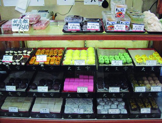 商店街公式ガイドに掲載された大文字の店頭の写真。ガラスケースの段に和菓子のトレーが並び、トレーの縁に「大文字」の文字が入っている。