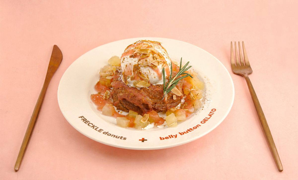 「FRECKLE donuts + belly button GELATO」と縁に印刷された白い皿。フレンチクルーラーの上にジェラートとスライスアーモンド、ローズマリー、角切りの果実が添えられている