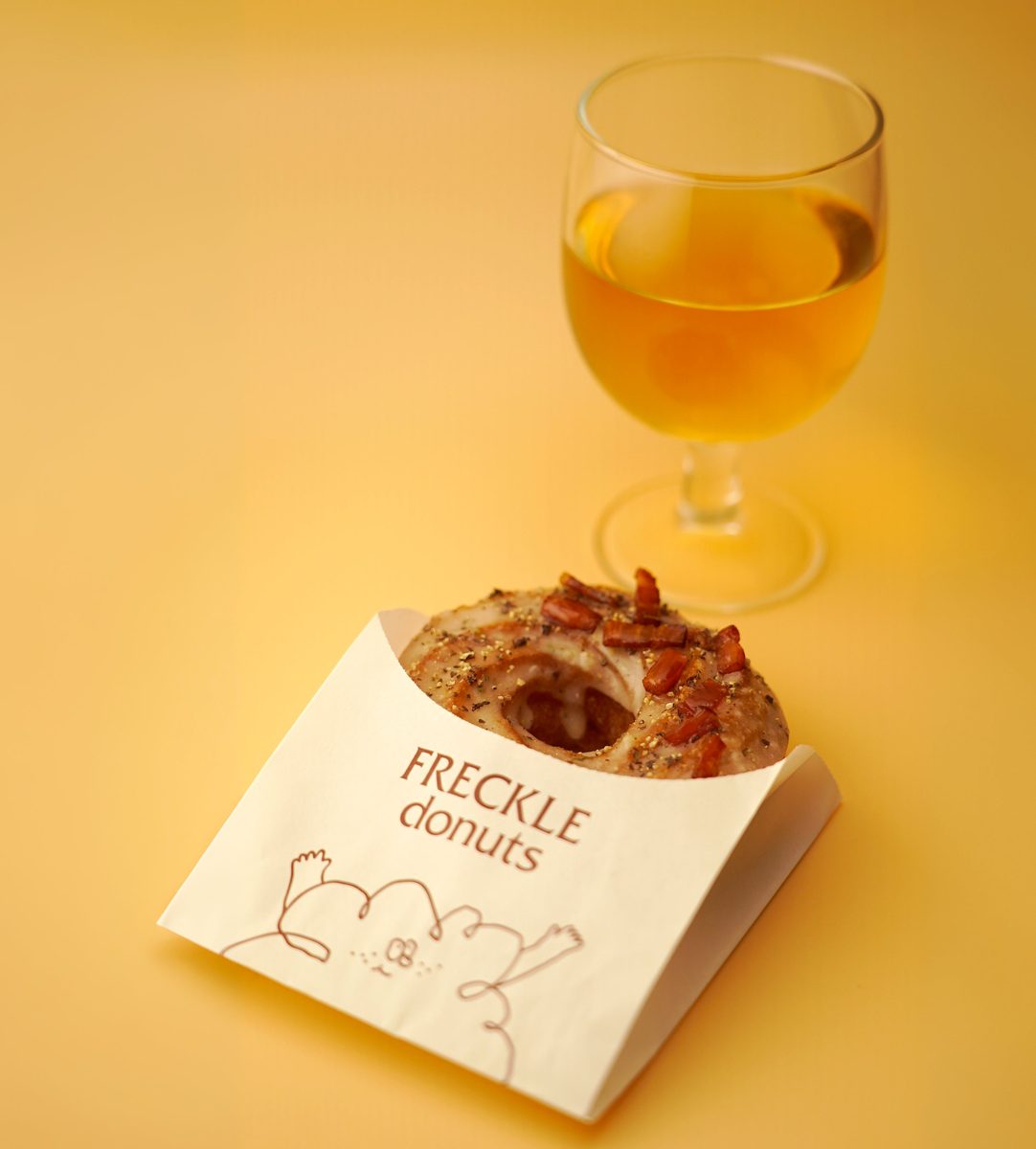 「FRECKLE donuts」と印刷された紙の包みに入った、ベーコンと黒こしょうをのせたフレンチクルーラー。奥に琥珀色の液体が入ったワイングラス