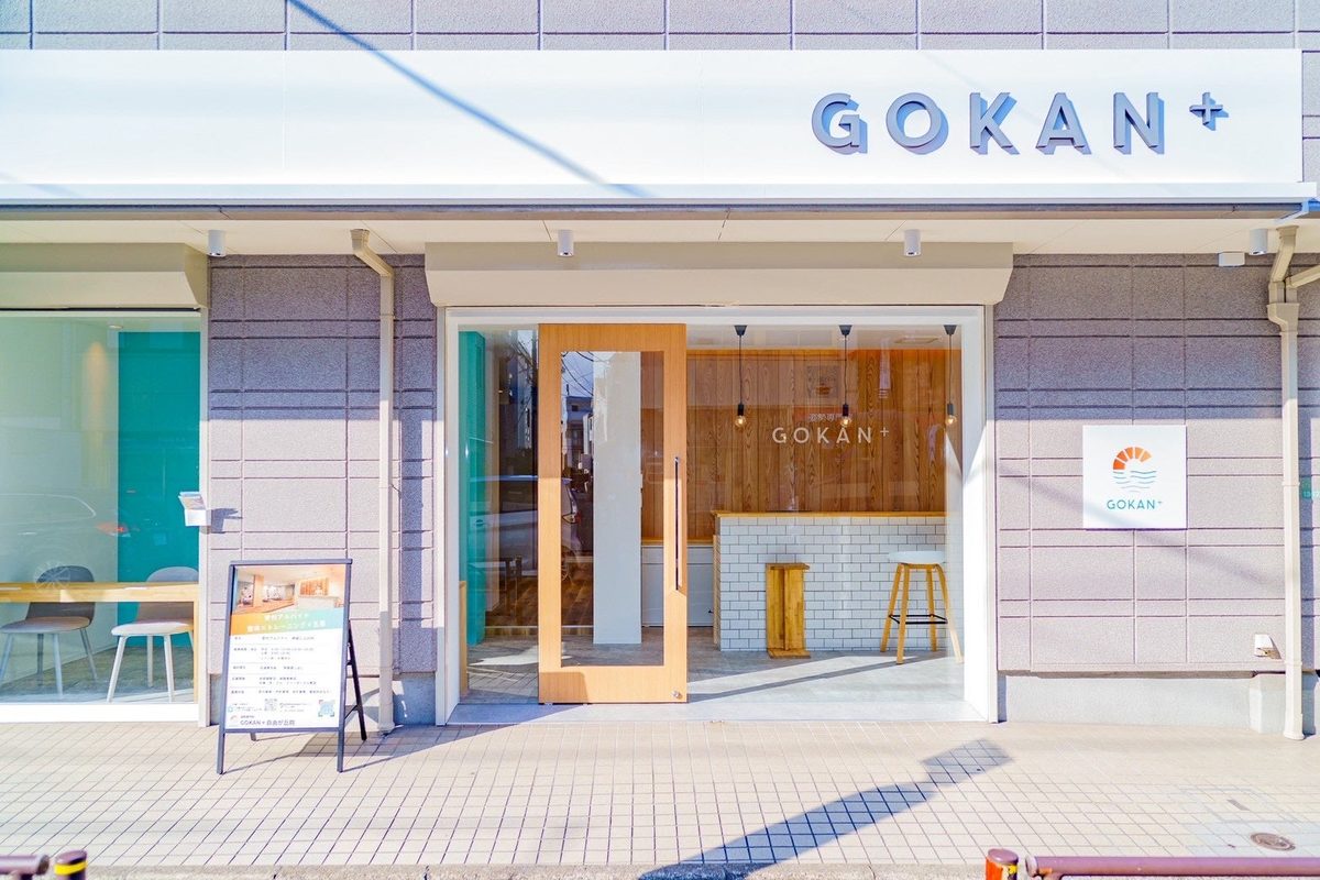 白いひさしに「GOKAN +」と掲げられた店舗の正面。開け放たれた木の扉の奥に、白タイルのカウンターと木のスツールが見える