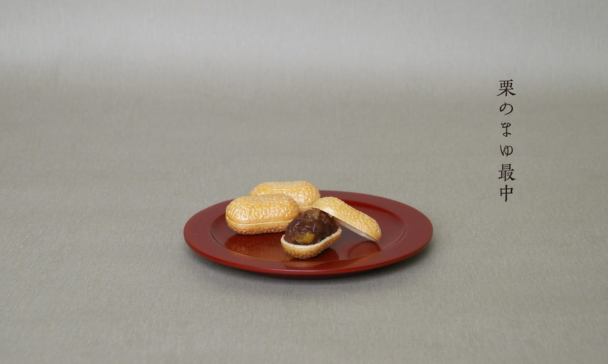 朱塗りの丸皿にのった、繭の形をした最中が3つ。手前の1つは割られ、栗の入った餡が見えている