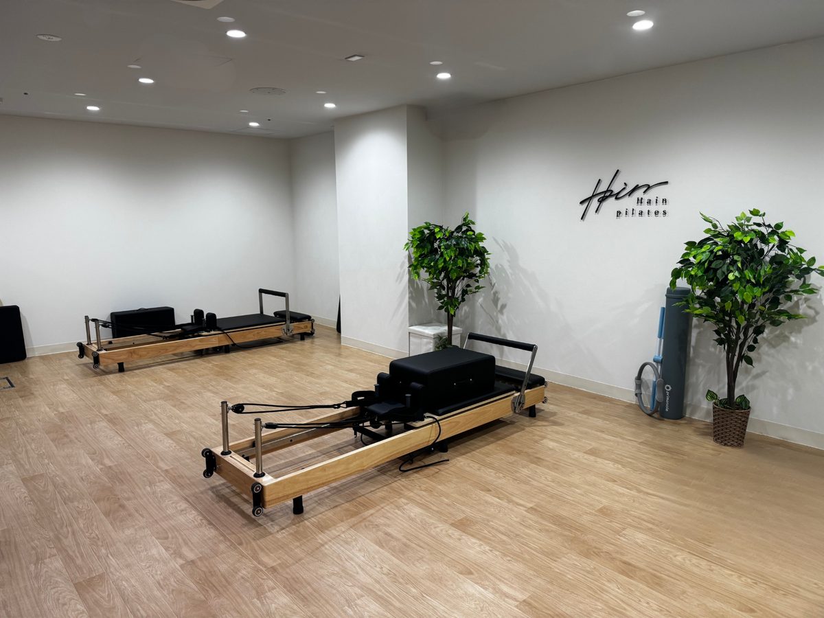 Hain pilates 自由が丘店のスタジオ内観。リフォーマー2台と Hain pilates のロゴが入った壁、観葉植物、木質フロアが写る