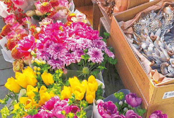 木の台に並べられた花束。手前に黄色いチューリップ、中央にピンクや紫の菊とバラ、右手の木箱にドライフラワーの束が立てられている