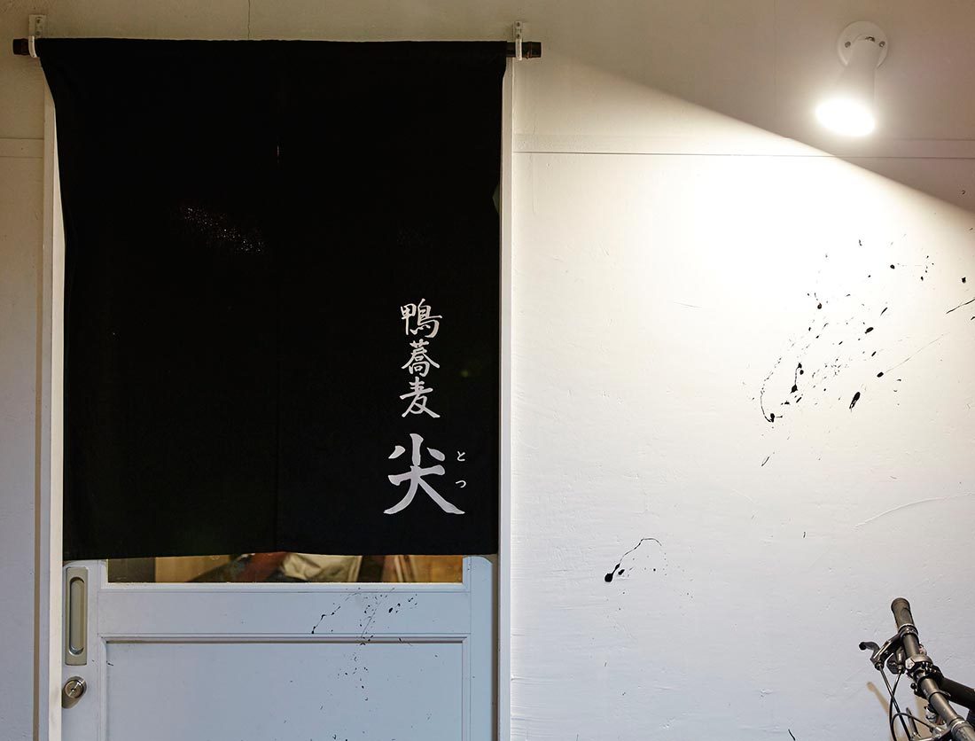 鴨蕎麦 尖の公式サイトで公開された写真。白い壁の入口に、「鴨蕎麦 尖」と白い文字が入った黒いのれんが掛かり、その下に白い扉、右上に照明、右下に自転車のハンドルが写っている