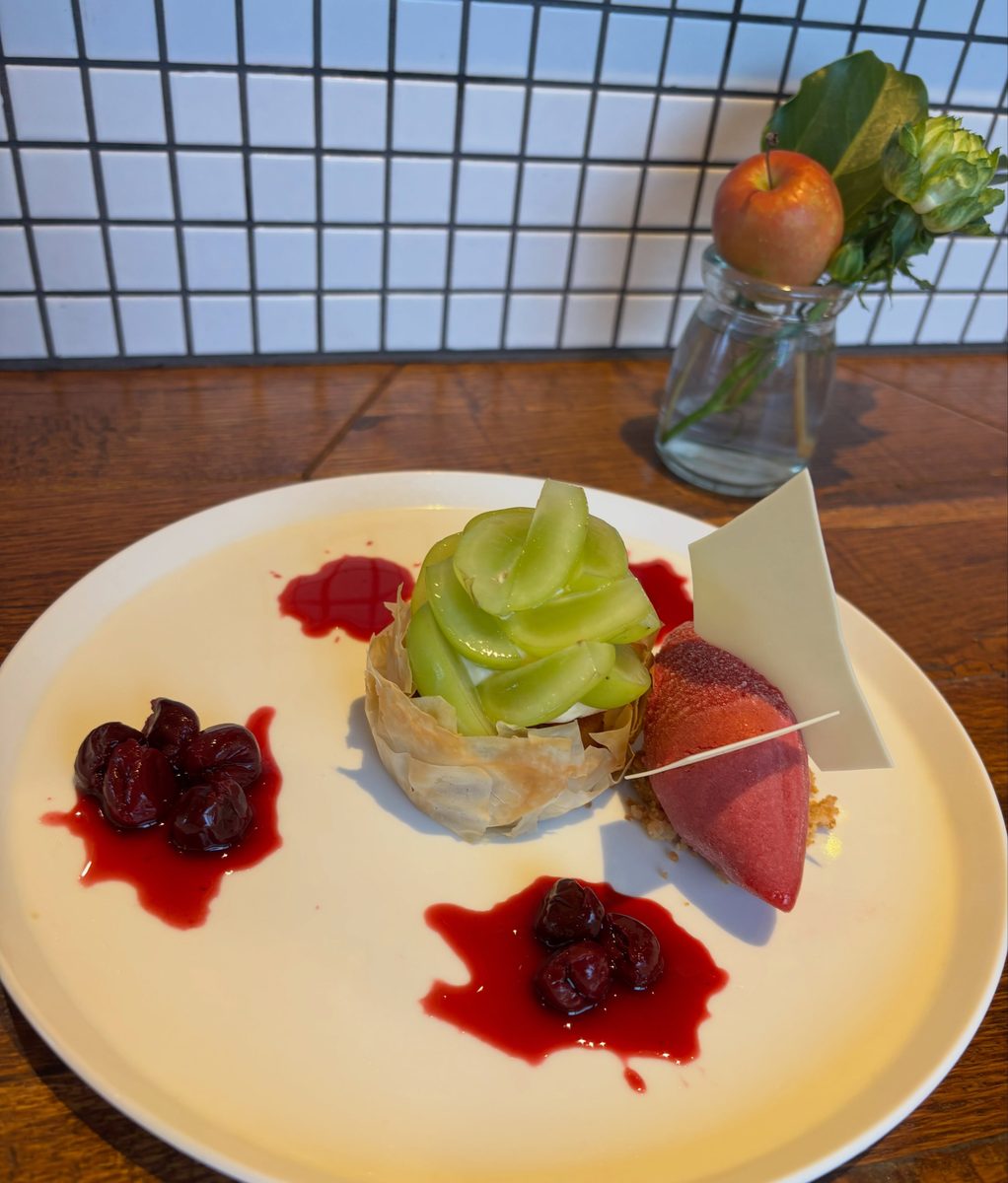 白い大皿に、緑色のクリームを絞った焼き菓子、赤いソルベ、赤い果実と赤いたれが盛られた一皿。奥は白いタイルの壁で、果実の付いた枝を挿した小瓶が置かれている。