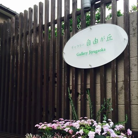 木の格子塀に掛けられた楕円形の白い看板。緑の文字で「ギャラリー 自由が丘」「Gallery Jiyugaoka」と書かれ、足元に花が植えられている。