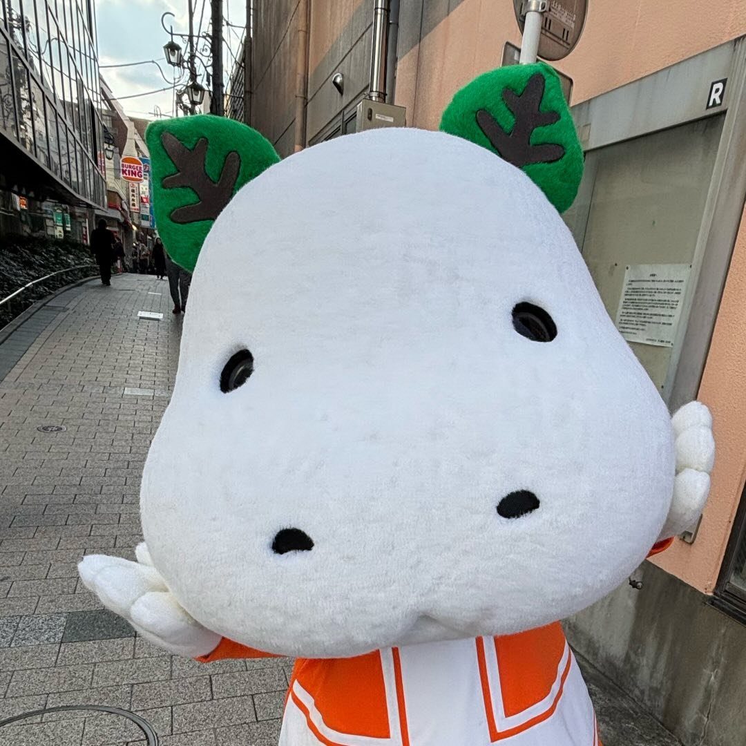 通りに立つキャラクター「しらかばくん」の着ぐるみ。白い顔に緑の葉の耳、オレンジの服