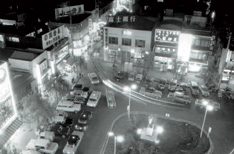 夜の駅前ロータリーを高い位置から写した白黒写真。円形の島の周りを車が取り囲み、奥のビルに「富士銀行」の看板が光っている