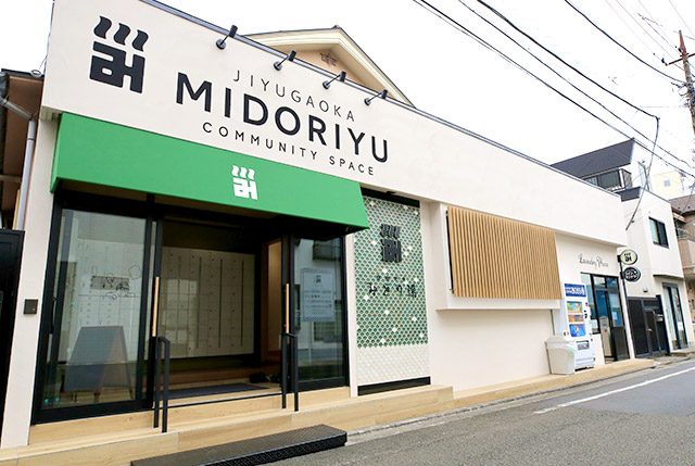 白い壁の建物の正面。緑の日よけの上に「JIYUGAOKA MIDORIYU COMMUNITY SPACE」の文字と湯のマーク、右手に「みどり湯」と読めるタイル貼りの看板がある