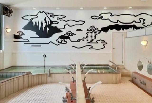 銭湯の浴場。白い壁面いっぱいに富士山と雲、松を黒い線で描いた絵があり、その下に緑がかった湯をたたえた浴槽が並ぶ