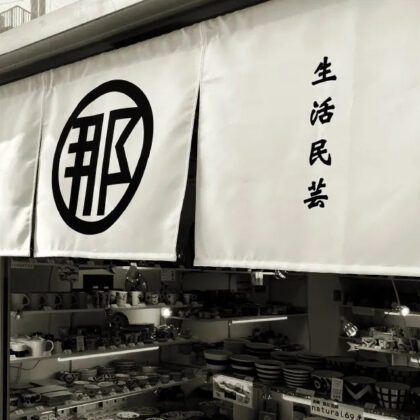 自由が丘商店街の公式ガイドの生活民芸 なかむた紹介ページで公開された白黒写真。丸で囲んだ「那」の字の印と「生活民芸」の文字が入った白いのれんが店先に下がり、その奥の棚に器が並ぶ