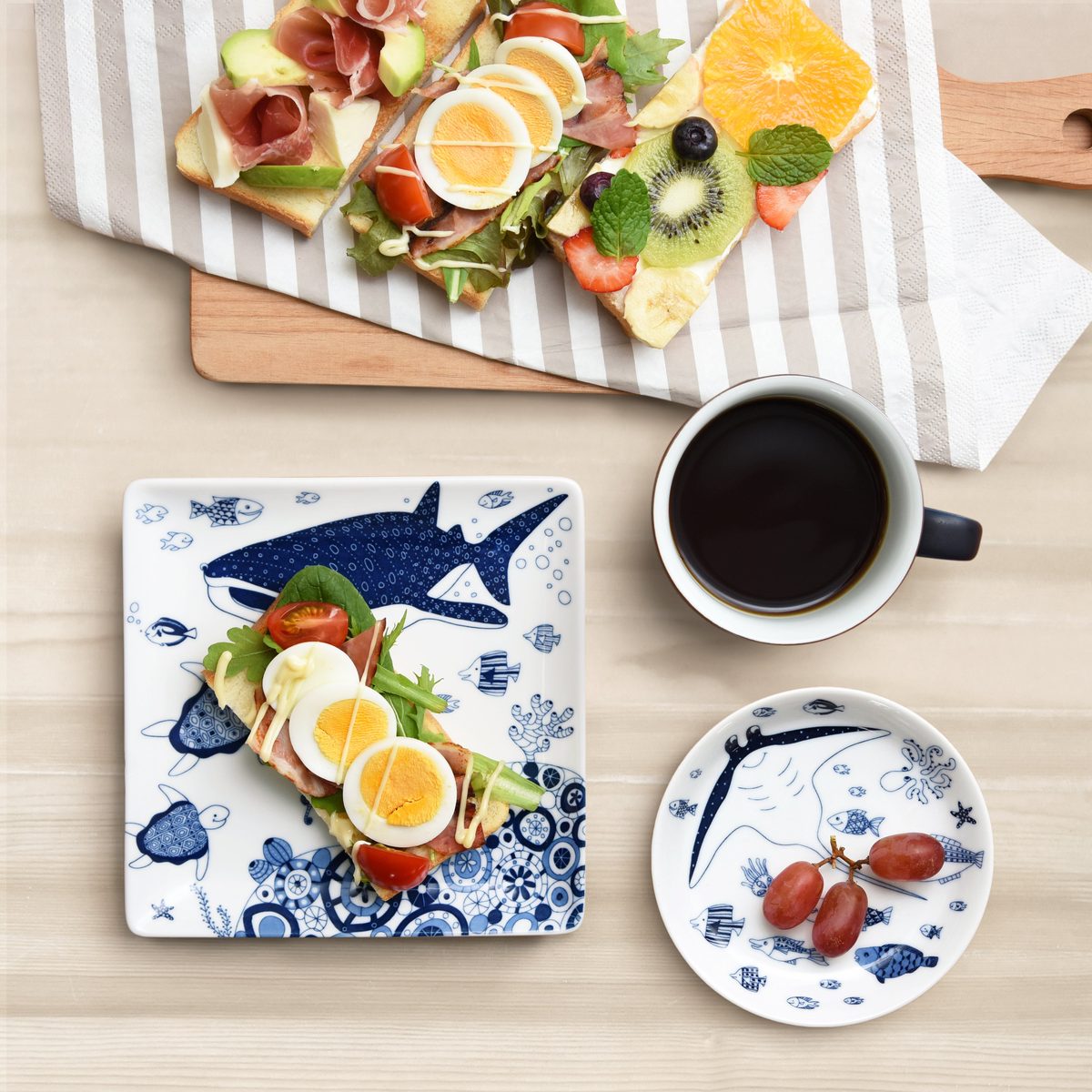 上から撮った食卓。サメや亀、魚の絵柄を藍色で描いた白い角皿にゆで卵と野菜をのせたパン、同じ絵柄の丸い小皿にぶどう、黒いコーヒーの入ったカップ、奥の木のボードに具をのせたパンが並ぶ