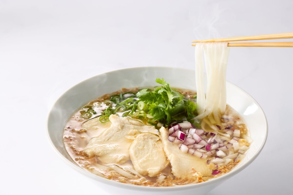白い器に張られた澄んだ出汁の麺料理。箸で平たい白い麺を持ち上げ、湯気が立つ。蒸した鶏肉のスライス、パクチー、小口ねぎ、角切りの紫玉ねぎがのる
