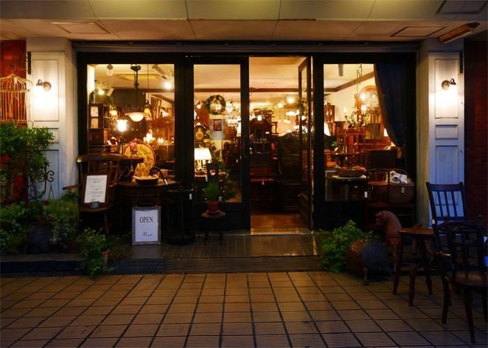ガラス扉の向こうに照明の灯った店内と木製家具が見える店舗入口。手前はタイル敷きで、扉の左右に壁付けの照明と鉢植えが置かれている。