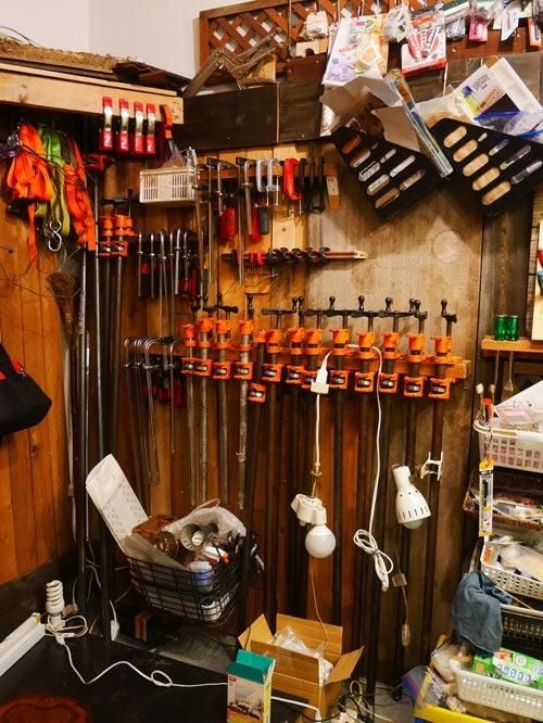 板張りの壁一面に、オレンジ色のクランプや工具が掛けられた作業場の一角。