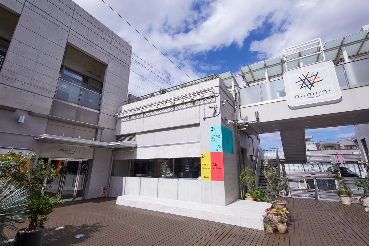岡田不動産の開業リリースで公開された建物の画像。木のデッキの奥にコンクリートの建物があり、張り出した看板に「mimimi JIYUGAOKA」、壁面に「2F EAT Restaurant / ART Art Space」「3F SEED Farm」と書かれている